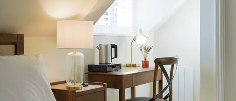 A hotel room with a bed, a desk with a lamp, a kettle, and a chair.