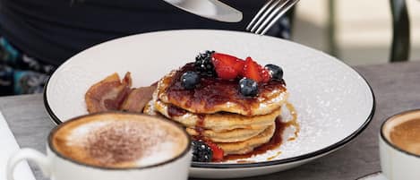 Eine Person isst Pfannkuchen mit Beeren und Sirup, eine Tasse Kaffee und ein Teller Essen.