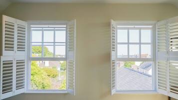 Two windows with white shutters, one with a view of greenery and another with a view of rooftops.