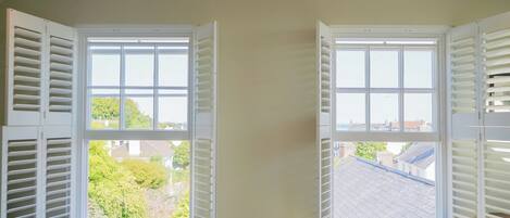 Two windows with white shutters, one with a view of greenery and another with a view of rooftops.