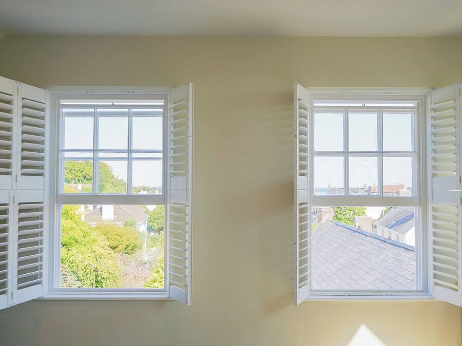 Two windows with white shutters, one with a view of greenery and another with a view of rooftops.