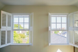Two windows with white shutters, one with a view of greenery and another with a view of rooftops.