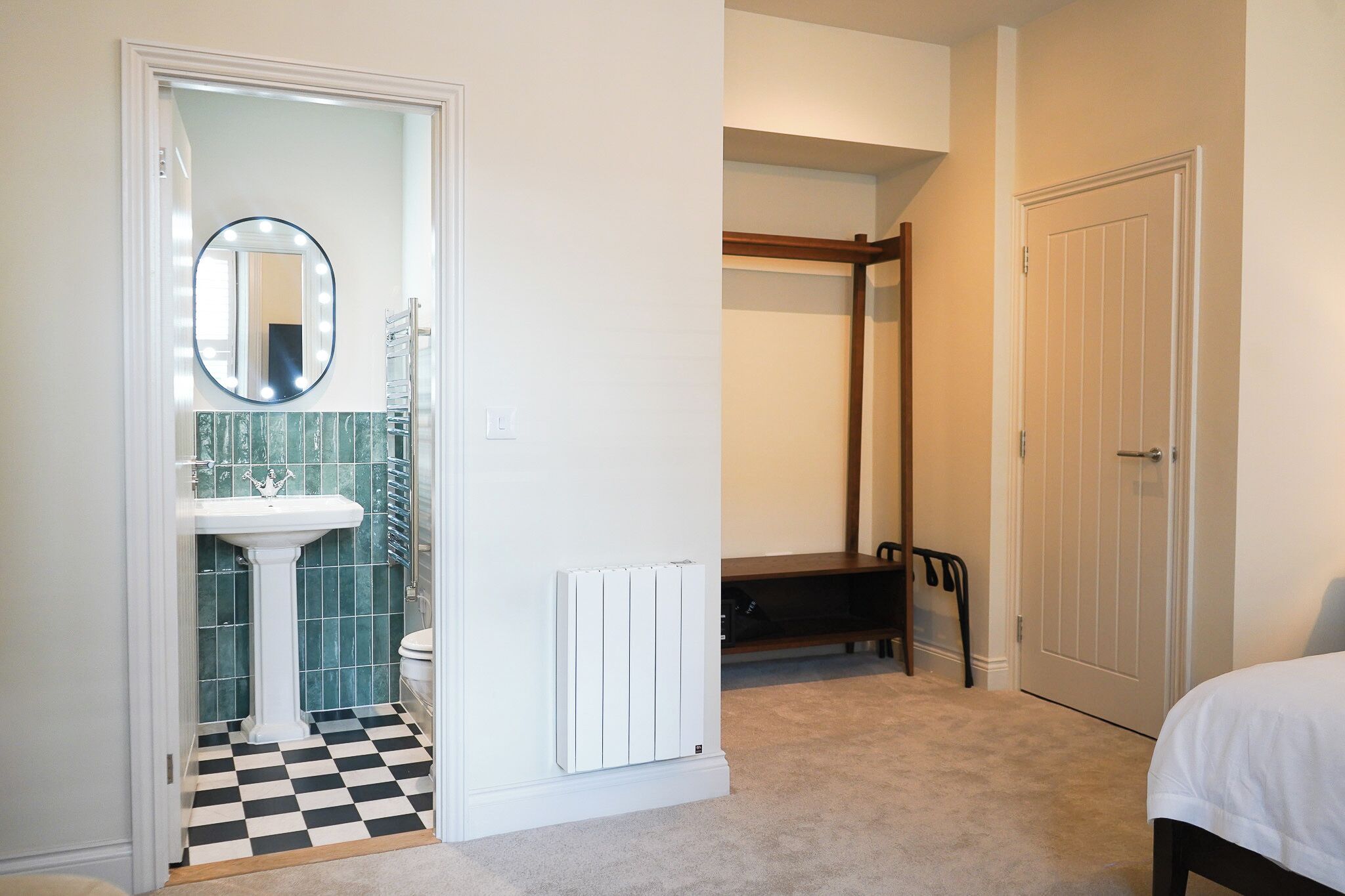 A bedroom with a bed, a white door, a radiator, a wooden chair, and a bathroom with a sink and mirror.