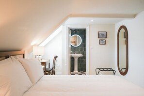 Hotelkamer met een bed, bureau, stoel, ronde spiegel en een badkamer met wastafel en betegelde wand.