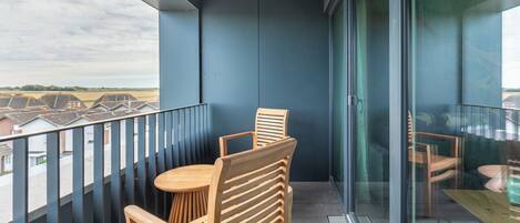 Un balcón con una mesa y sillas de madera, una puerta de cristal y vistas a casas a lo lejos.