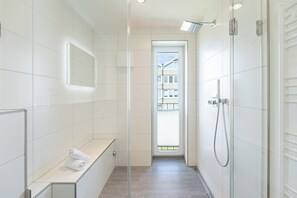 Ein modernes Badezimmer mit Duschkabine aus Glas, einer Sitzbank mit Handtüchern und einem Durchgang zur Außenwelt.
