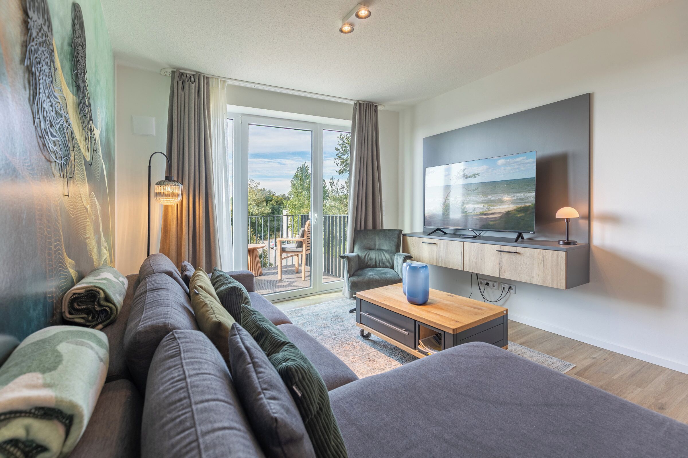 Een moderne woonkamer met een grote bank, een salontafel en een flatscreen televisie aan de muur bevestigd. Er is een schuifdeur naar een buitenruimte met uitzicht op de lucht en bomen.
