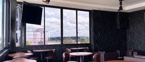A restaurant with round tables and chairs, a flat-screen TV mounted on the wall, and large windows offering a view of the outside.
