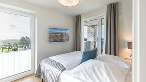 Camera d'hotel con una grande finestra, un letto con lenzuola bianche, un cuscino blu, un quadro appeso al muro e un balcone con vista.