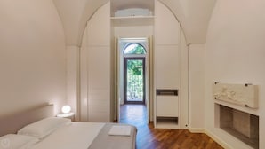 Une chambre à coucher comprenant un lit, une table de chevet, un miroir, une cheminée et une porte en arc donnant sur un balcon.
