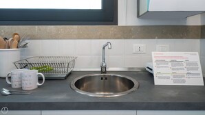 Un évier de cuisine en acier inoxydable, doté d’un robinet moderne, d’un égouttoir à vaisselle en métal et de deux tasses blanches arborant la marque Italianway. Un panneau d’information sur le recyclage est visible sur le mur.