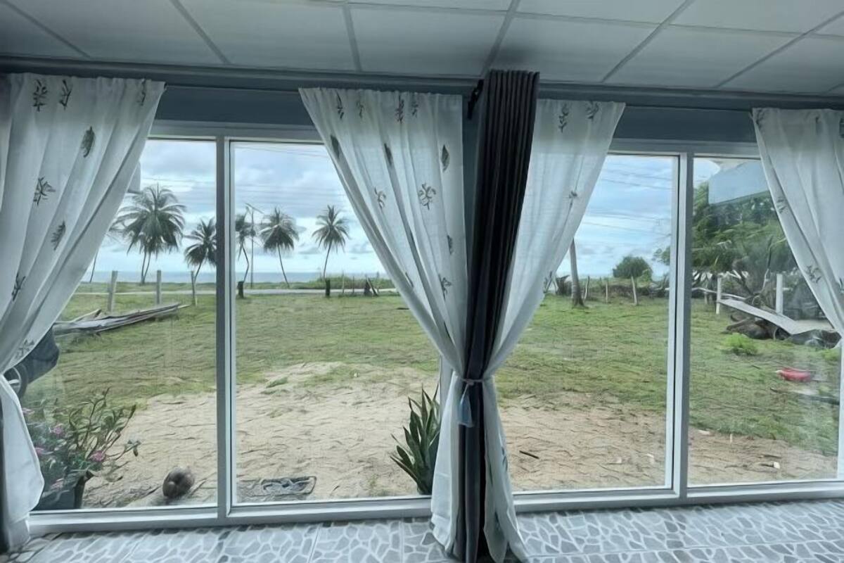 Ein Zimmer mit großen Fenstern, Vorhängen und Blick auf eine Rasenfläche mit Palmen.