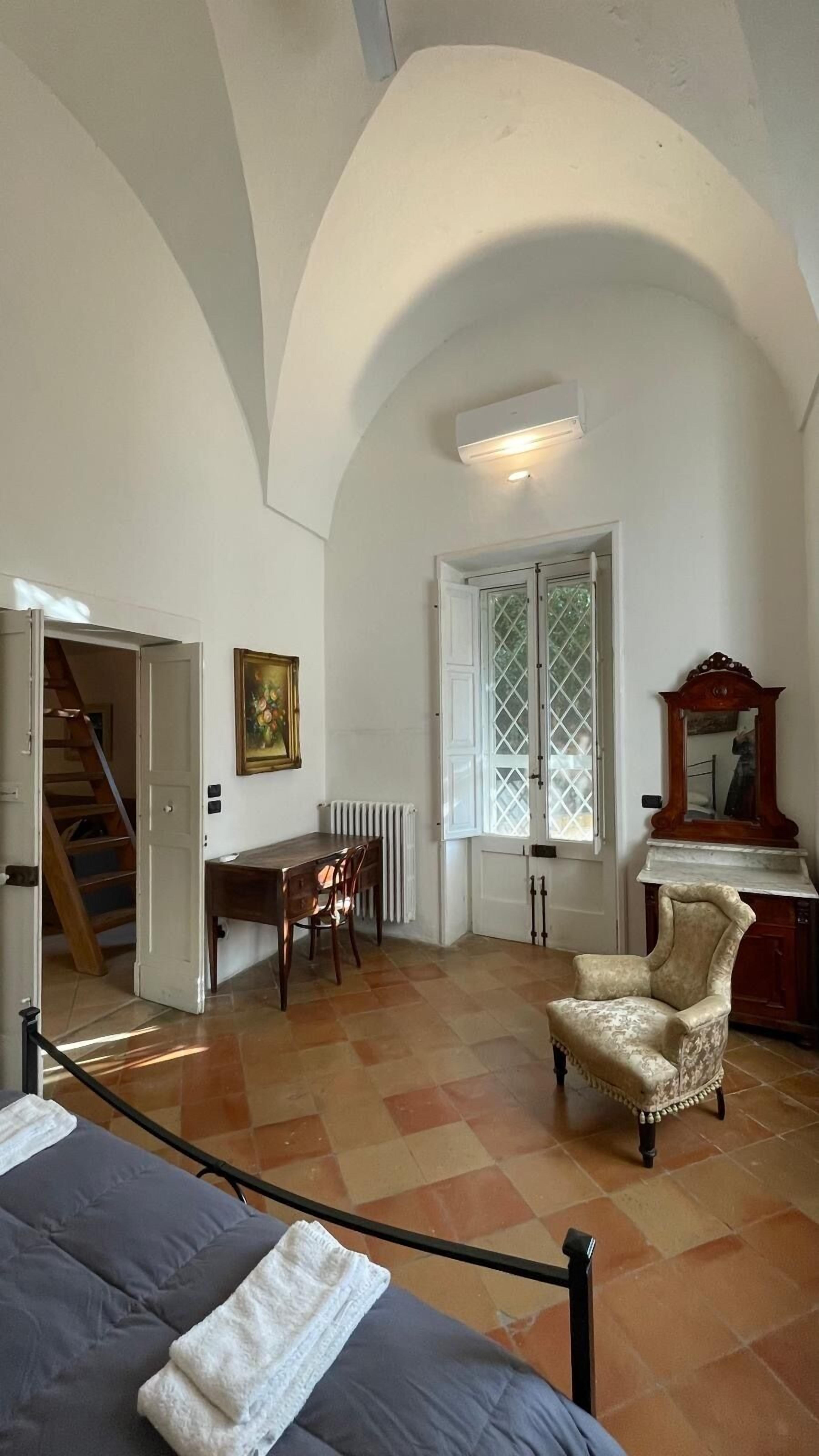 Ein Zimmer mit einer hohen, gewölbten Decke, einem Esstisch mit Stühlen, einem Heizkörper, einem großen Fenster mit Gitterwerk und einem Bett mit Kopfteil.