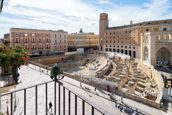 Ein antikes römisches Amphitheater mit Steinsitzen und einer zentralen Bühne, umgeben von modernen Gebäuden.