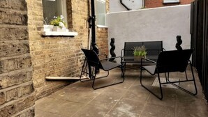 A patio with two black metal chairs and a table, a brick wall, and a small white shelf with plants. - Big Family Home! FREE Parking, Fast WiFi, Garden (Kingston upon Thames)