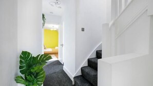 A hallway with a staircase, a plant, and a door leading to a room with a yellow wall. - Big Family Home! FREE Parking, Fast WiFi, Garden (Kingston upon Thames)