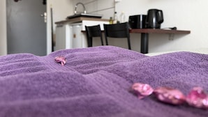 A room with a bed covered in a purple blanket, a small pink toy on the bed, a kitchen area with a microwave, and a television mounted on the wall.