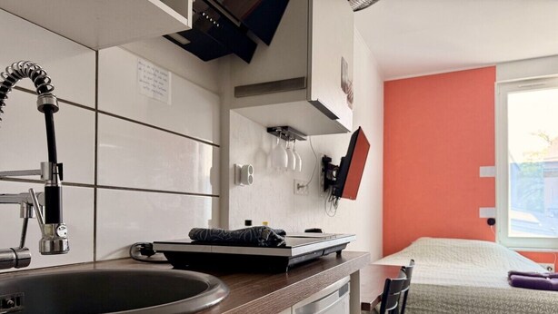 A compact kitchen with a sink, a stove, and a microwave. There is a bed with a white and orange wall, and a window providing natural light.