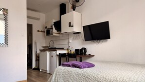 A compact hotel room with a bed, a desk, a kitchenette, and a TV mounted on the wall.