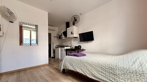 A bedroom with a bed, a desk, a chair, a mirror, and a kitchenette.
