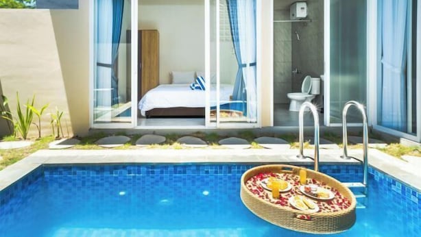 Habitación de hotel moderna con puerta de vidrio que da a un balcón, baño privado y zona de piscina con una cesta de comida flotando.