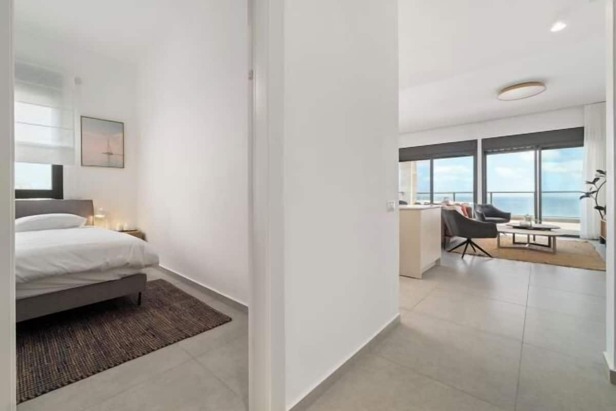 A modern hotel room with a bed, bedside table, and a desk with a chair. There is a large window offering a view of the sea.