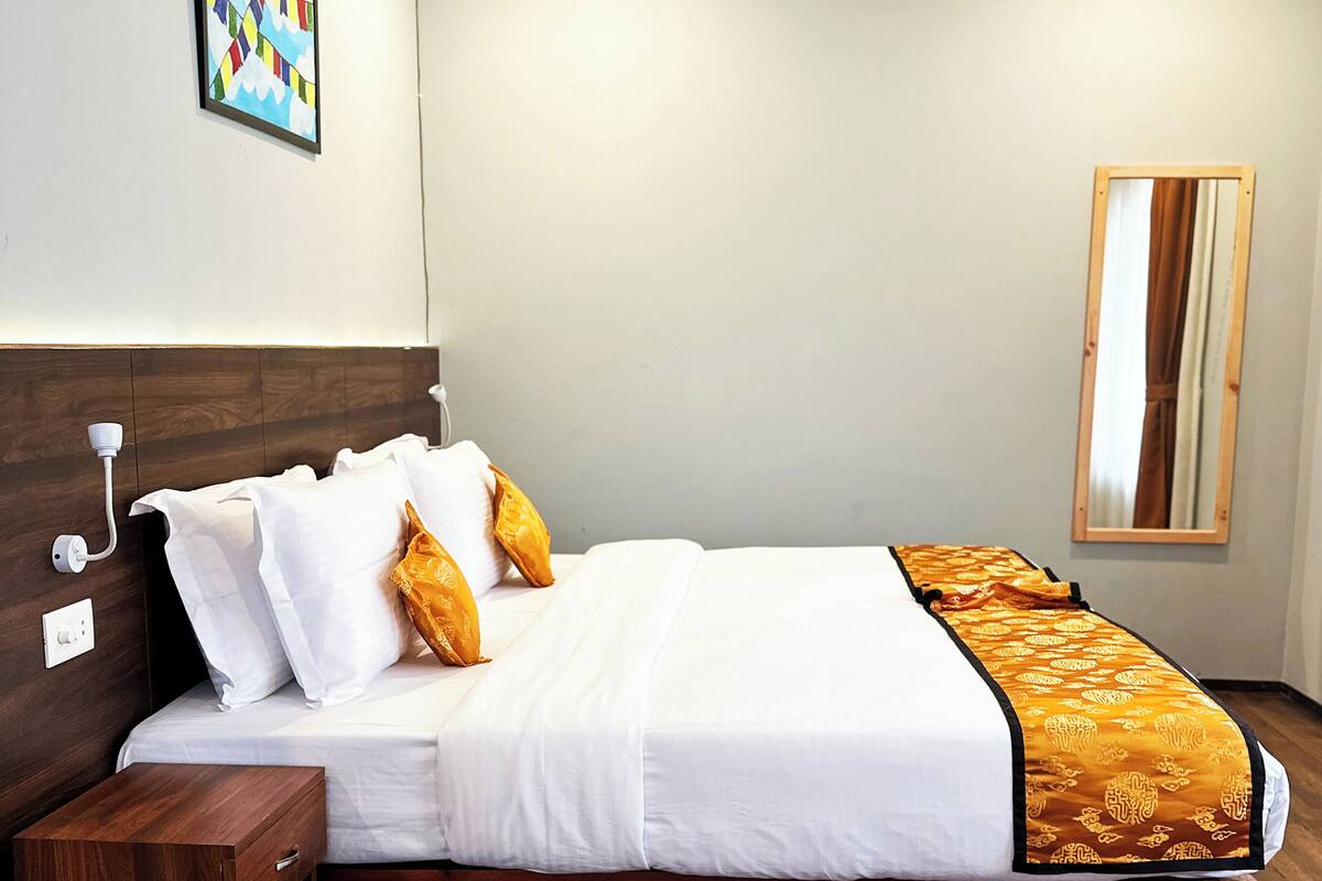 A neatly made bed with white linens and two orange pillows, a wooden headboard, a bedside table with a drawer, and a mirror on the wall.