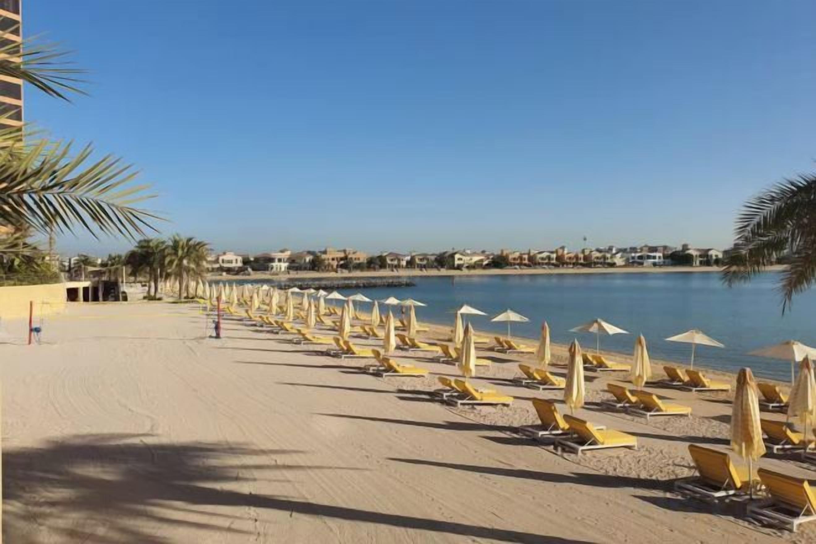 Une plage bordée de rangées de transats et de parasols, un ciel dégagé et une mer calme.