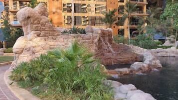 A landscaped area with a rock formation and a small water feature, surrounded by greenery and a building with balconies in the background.