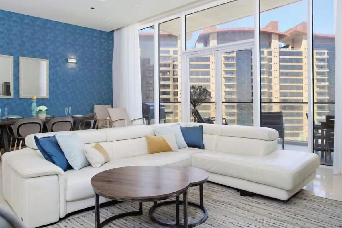 A modern living room with a white sectional sofa, a round coffee table, and large windows offering a view of high-rise buildings.