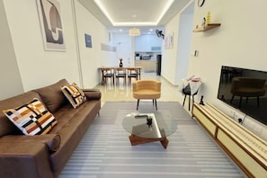 A modern living room with a sofa, armchair, coffee table, TV, and kitchen in the background.