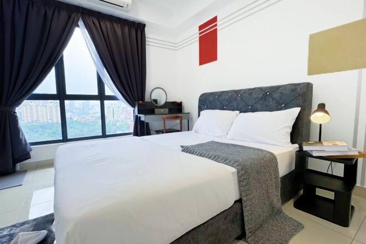 Habitación de hotel con una cama grande, una mesita de noche con lámpara, un escritorio pequeño con silla, y vista a la ciudad a través de un ventanal amplio.