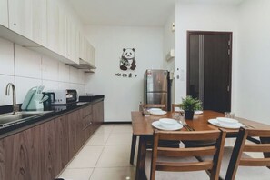 A compact kitchen with wooden cabinets, a stainless steel refrigerator, a microwave, and a coffee maker. There is a dining table set with plates and a potted plant. - Molek Little Joy Studio 4pax Bathtub with Balcony (Plentong)