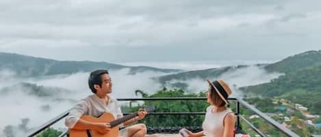 Two people sitting on a colorful blanket with a guitar, overlooking a valley with mist-covered hills.