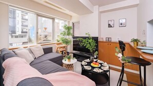 A modern living room with a sofa, a dining table with plates, a TV, and a kitchen area with cabinets and a chair. - SkyTree View - CoCoHouse Asakusa (Tokyo)