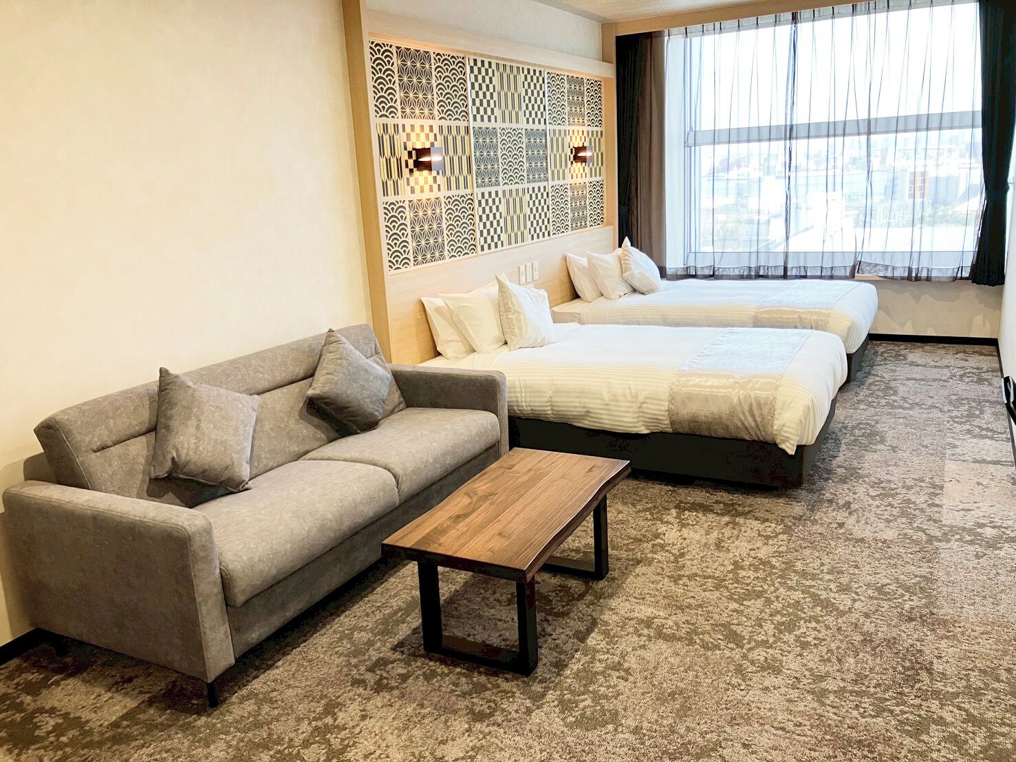 A hotel room with a sofa, a wooden coffee table, two beds, and a decorative wall panel.