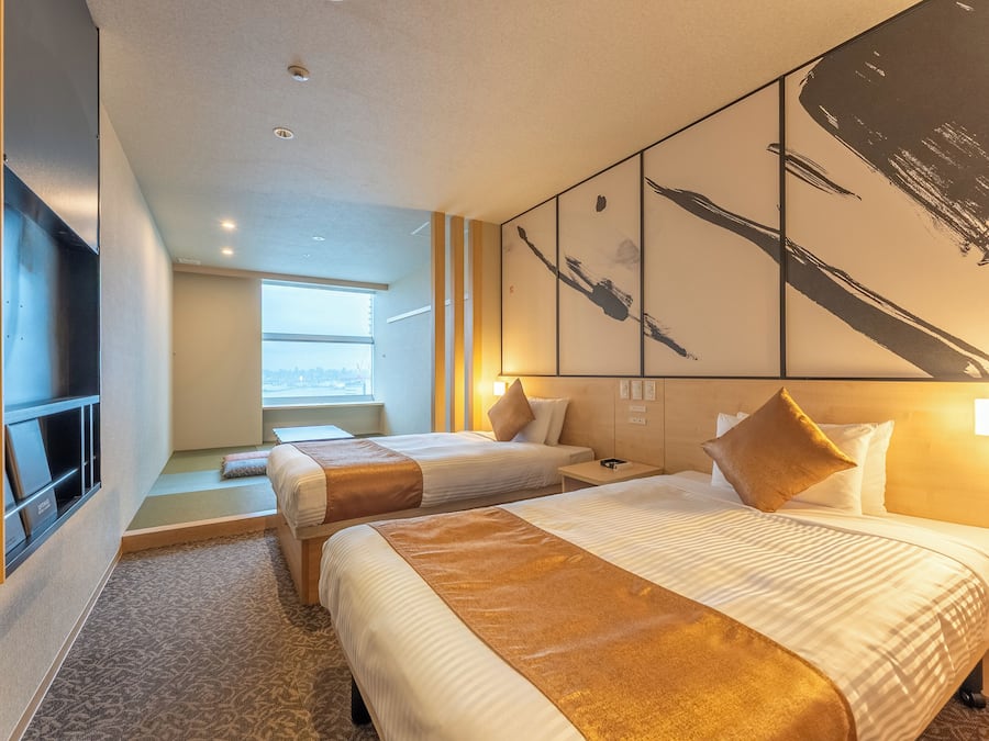 A hotel room with two beds, a large window, artwork on the wall, and a flat-screen TV.
