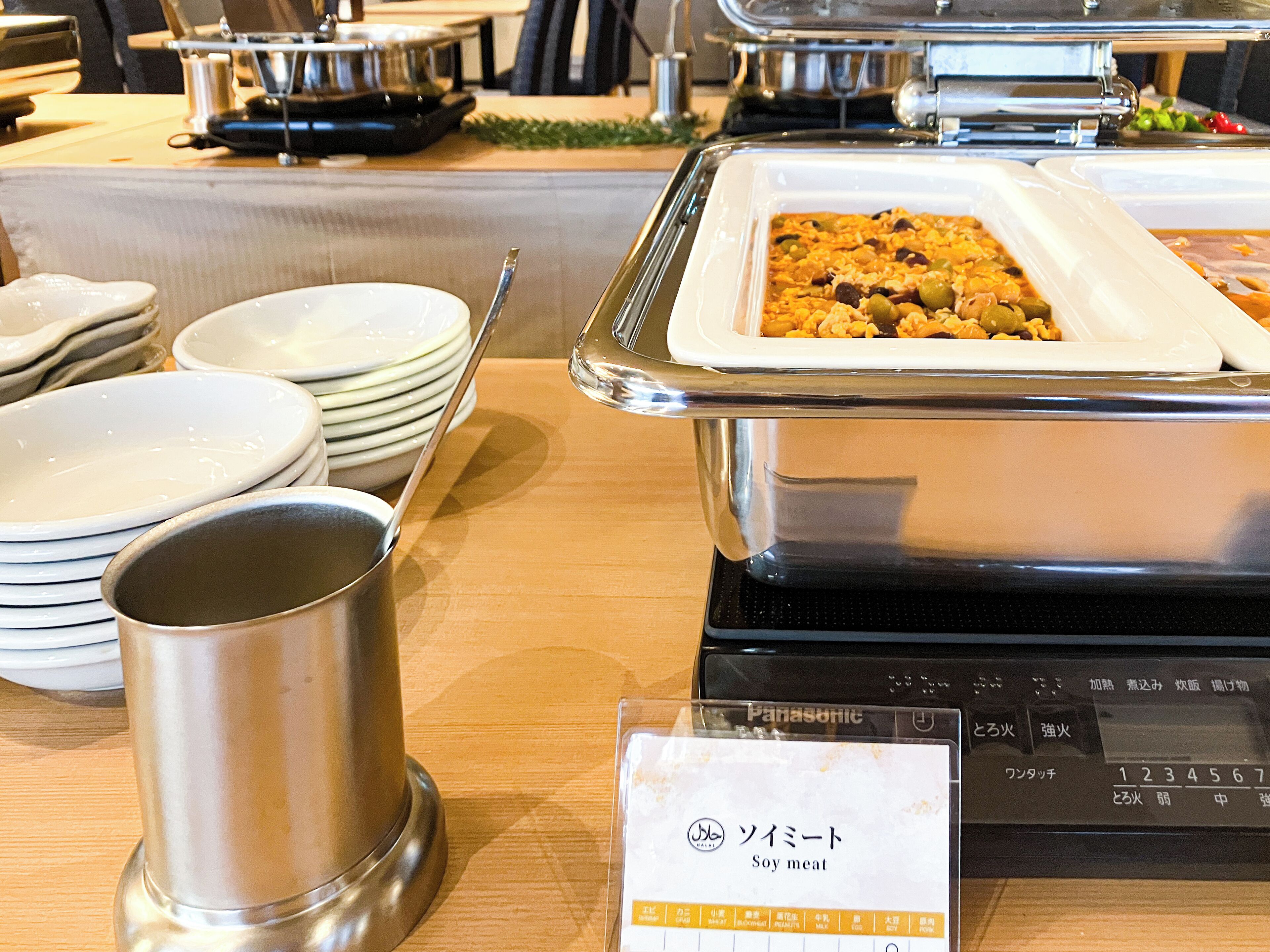A buffet setup with various dishes, including a labeled tray of soy meat, stacked plates, and a metal cup.