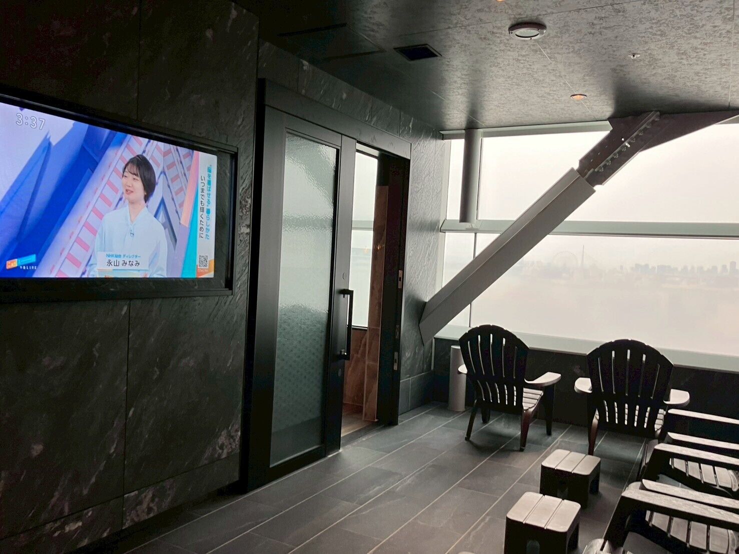Interior moderno com uma TV exibindo um telejornal, vista da cidade através de grandes janelas e um espaço de estar com cadeiras e mesas.