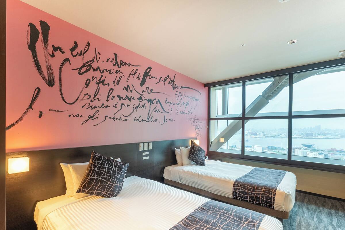 A hotel room with two beds, a large window with a city view, and a pink wall with handwritten text.