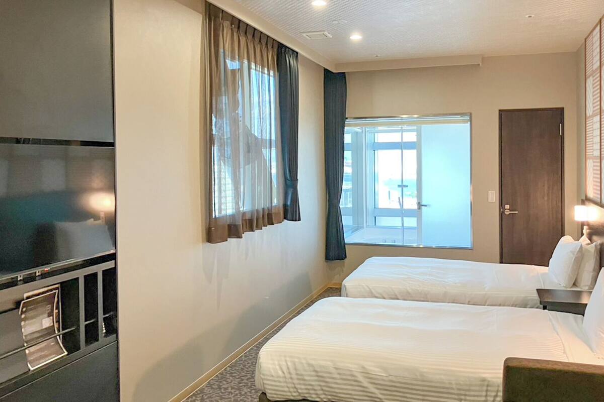 A hotel room with a bed, a TV, a window with curtains, and a door.