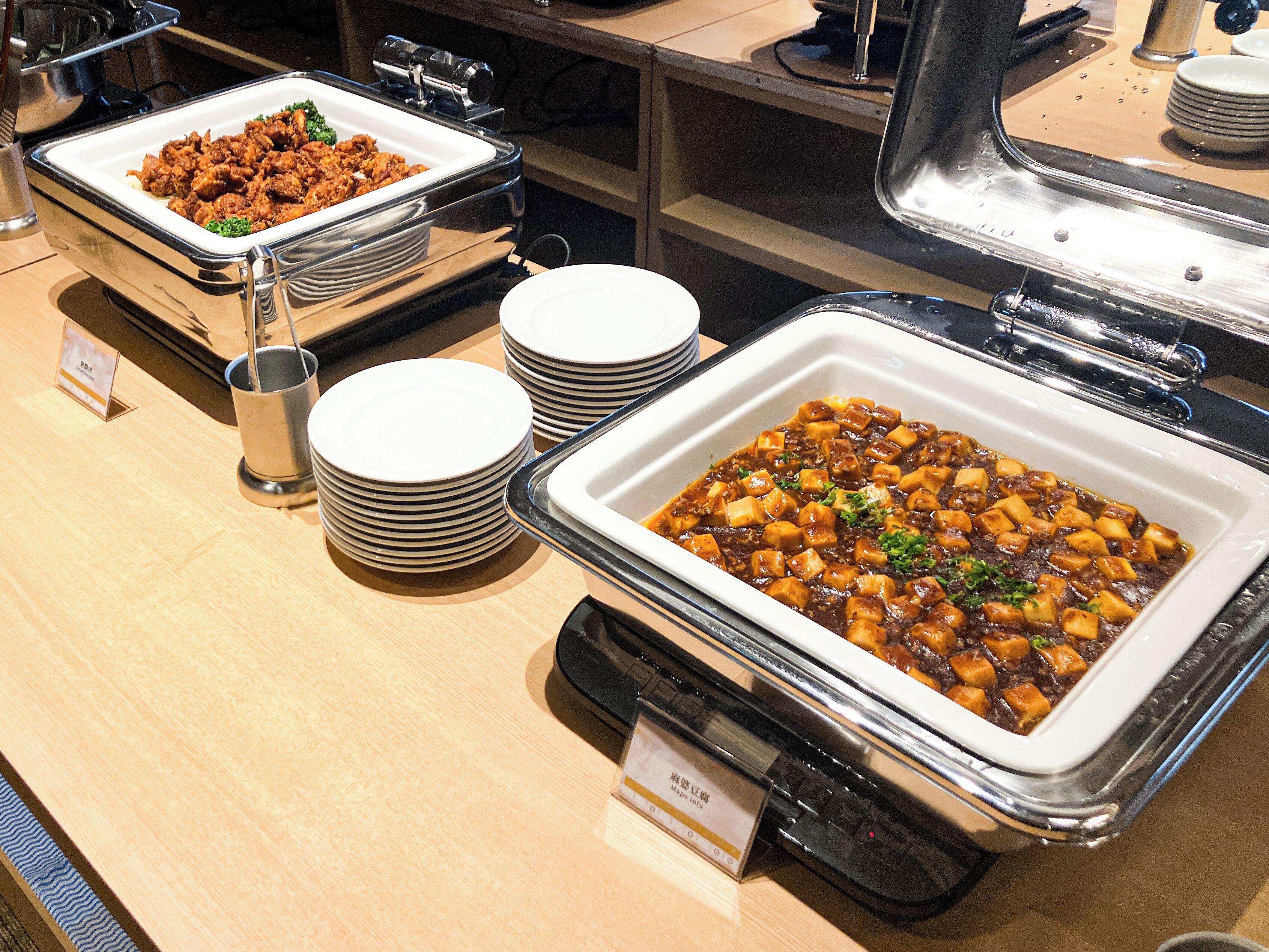 En buffet med flere retter, herunder en stor skål tofu i mørk sauce, omgivet af stablede hvide tallerkener og en metalbakke.