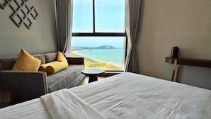 Une chambre d’hôtel avec un grand lit, un canapé avec des coussins, une petite table ronde et une vue sur la mer par une grande fenêtre.