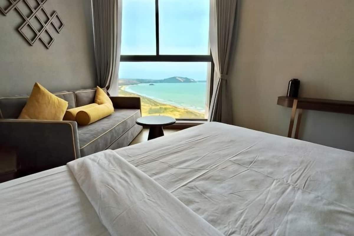 Une chambre d’hôtel avec un lit, un canapé avec des coussins, une petite table ronde et une vue sur la plage.