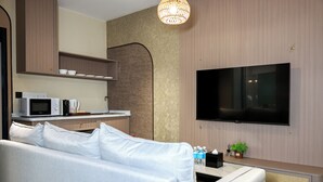 Un salon moderne avec un canapé blanc, un téléviseur à écran plat fixé au mur, un coin cuisine équipé d’un four à micro-ondes et d’une machine à café, ainsi qu’un luminaire décoratif.