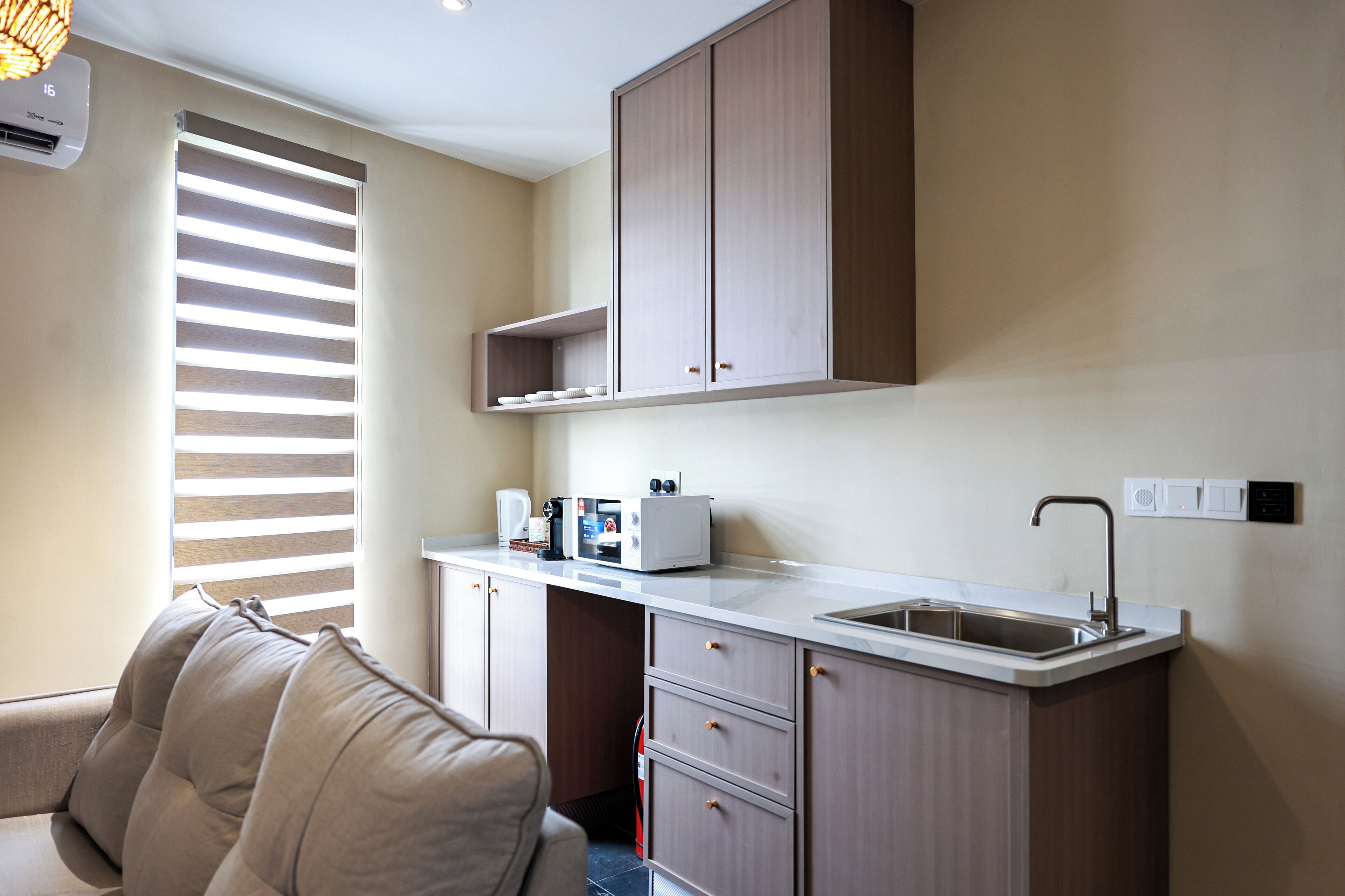 A compact kitchen with a built-in sink, a microwave, and a toaster. There is a window with blinds, a sofa, and a ceiling-mounted air conditioner.