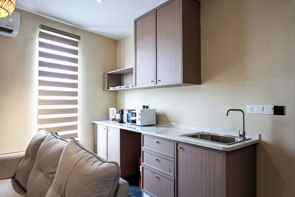 Cocina compacta con pileta integrada, microondas y tostadora. Hay una ventana con persianas, un sofá y un aire acondicionado instalado en el techo.