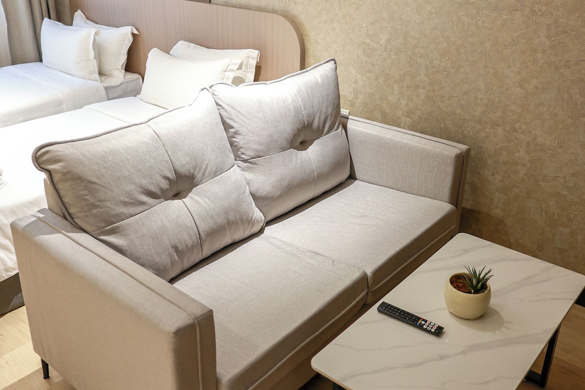 Une chambre d’hôtel moderne avec un canapé beige, une petite table sur laquelle est posée une plante en pot et un lit avec une literie blanche.