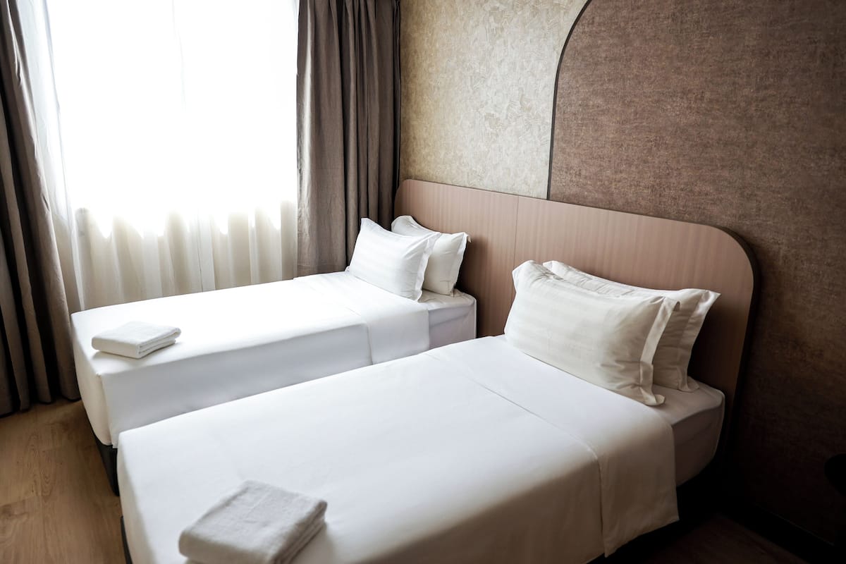 Une chambre d’hôtel avec deux lits simples, une grande fenêtre avec des rideaux et un mur à texture.