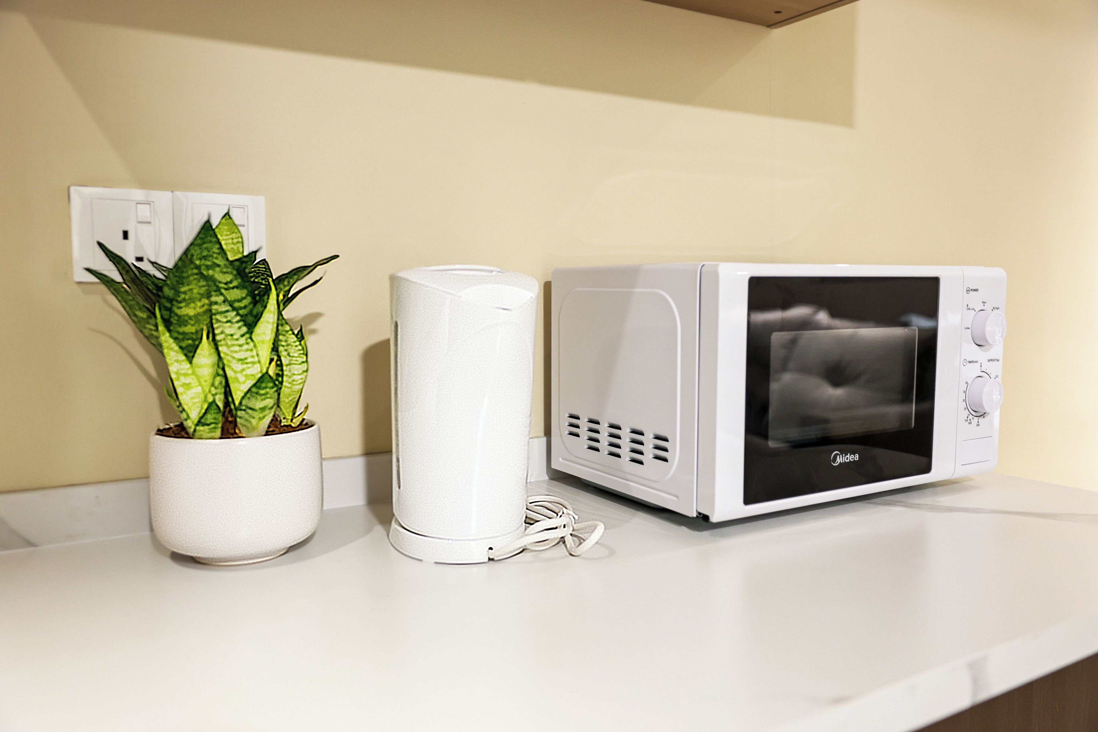 A microwave and a white cylindrical device on a white countertop, with a potted plant beside them.